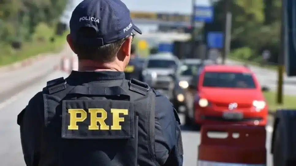 Agente da PRF de costas, com uniforme e boné. Ao fundo carros estão desfocados.