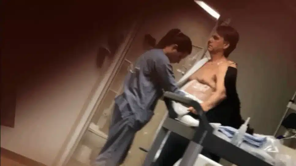 Carlos Bolsonaro divulgou uma foto em que o ex-presidente aparece em pé ao lado de uma cama hospitalar, com curativos na região abdominal, enquanto é auxiliado por uma profissional de saúde | Reprodução/X @CarlosBolsonaro - 16.mar.2026